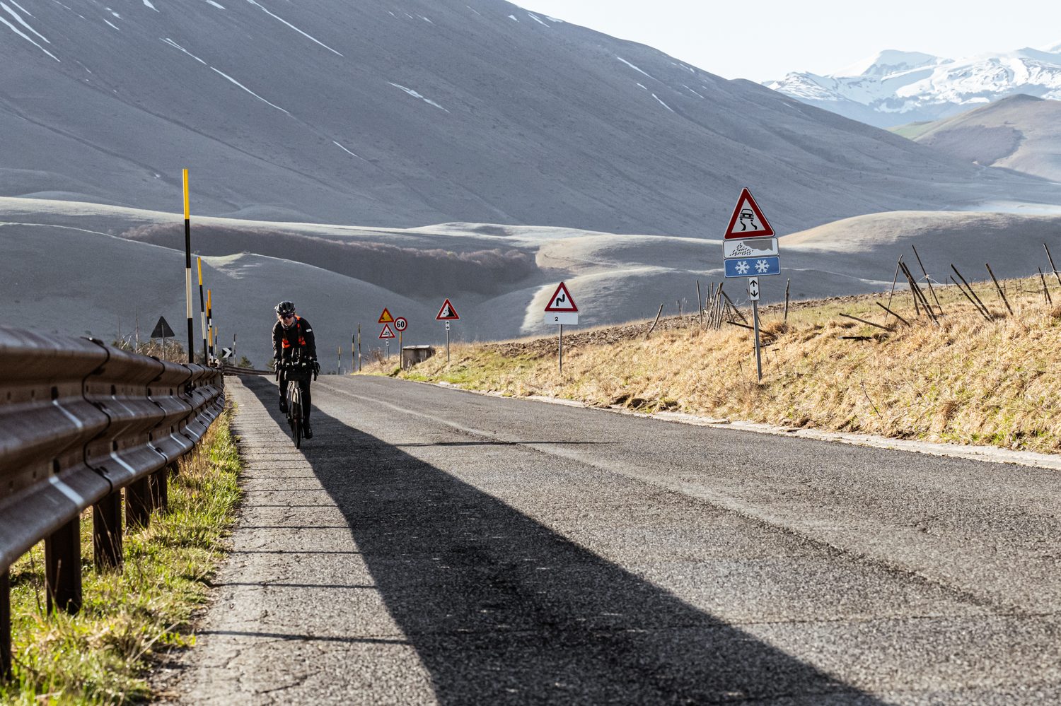 Rider climbing a mountain road past winter warning signs, snowy peaks behind, during TURN03 in Italy