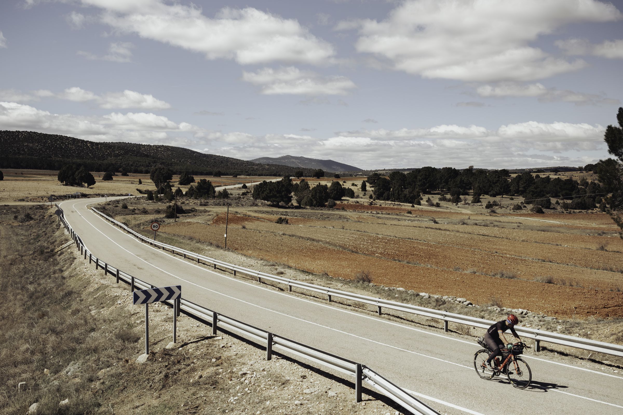 Rider climbing a quiet road through red fields and pine hills